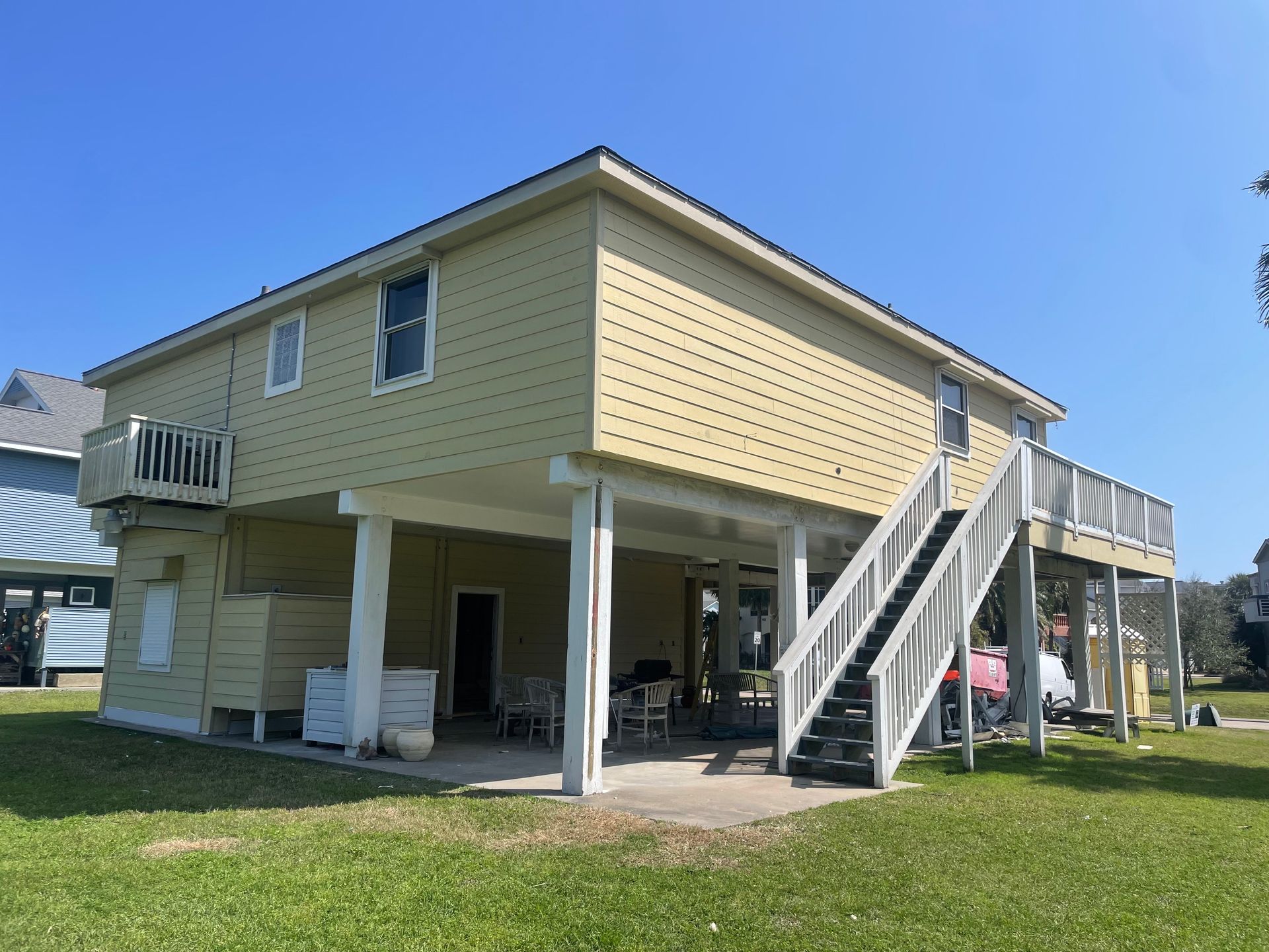 Two-story beige house on stilts. White deck and stairs lead up from the ground. Green grass and blue sky in the background.