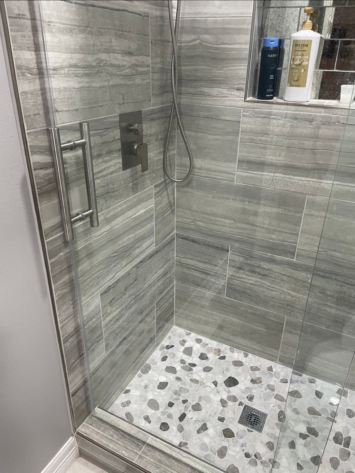 A modern shower with gray tile walls, a pebble tile floor, and a glass door. A shower handle and shower head are visible.