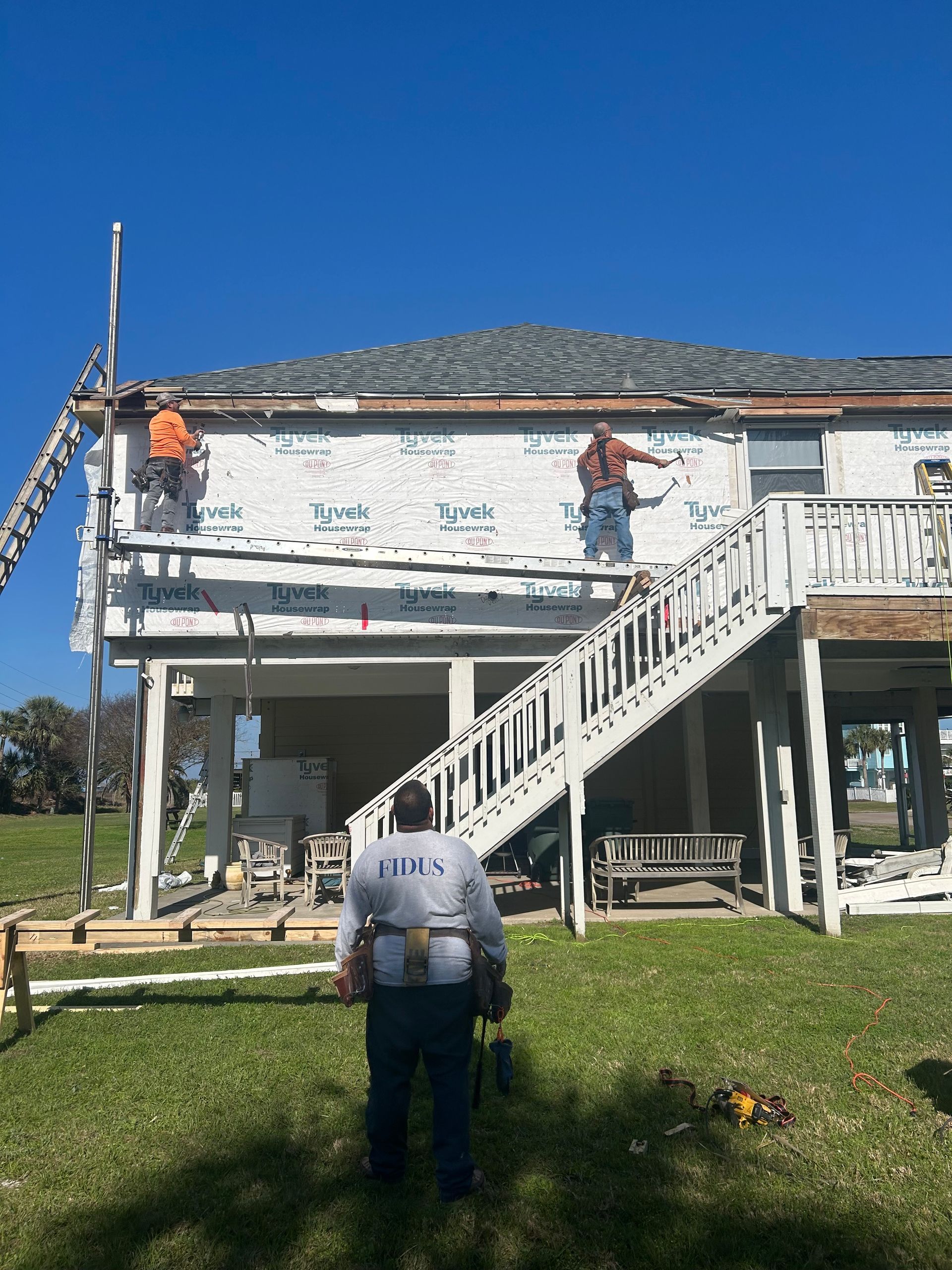Construction workers on a two-story house installing siding. The house is white with a staircase, and workers are on scaffolding.