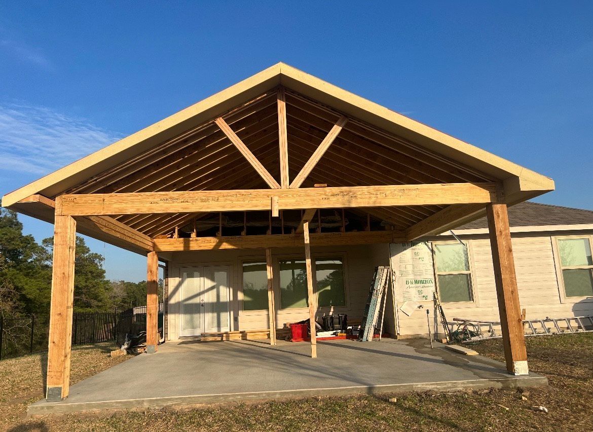 A partially built wooden patio cover attached to a house, over a concrete patio. Light blue sky.