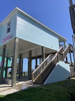 Blue beach house on stilts with wooden stairs. The house has a surfboard sign and is set on a grassy lawn.