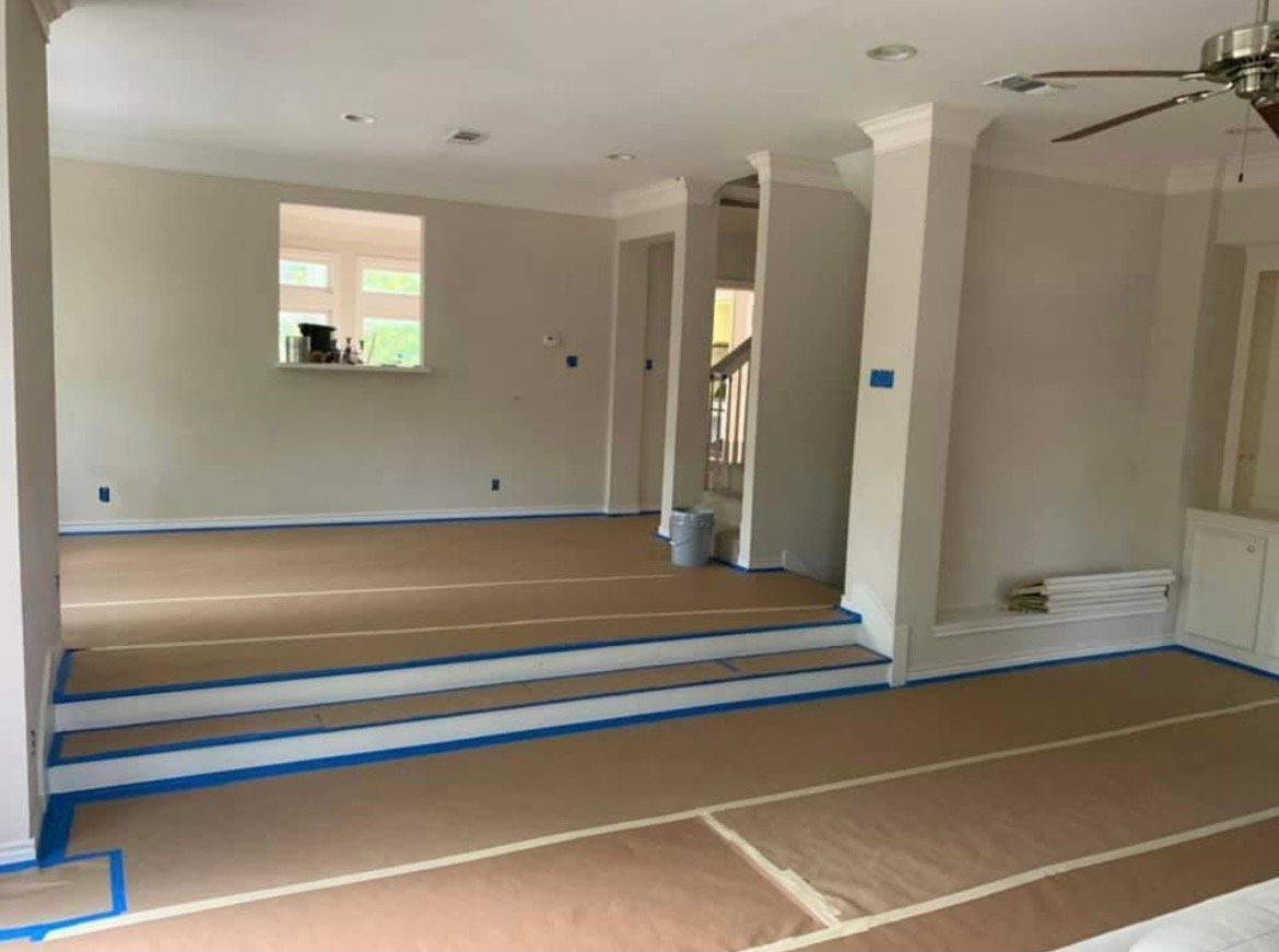An interior view of a room under renovation, with paint on walls, paper covering the floor, and blue tape lining the edges.