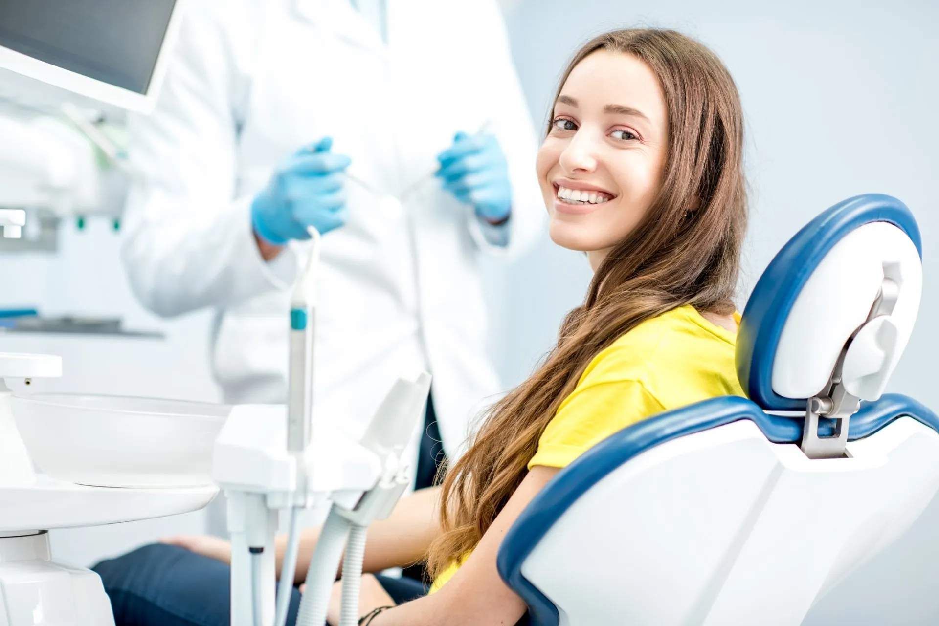 Woman in dental chair smiles, dentist in background. Yellow shirt, blue chair, dental equipment.