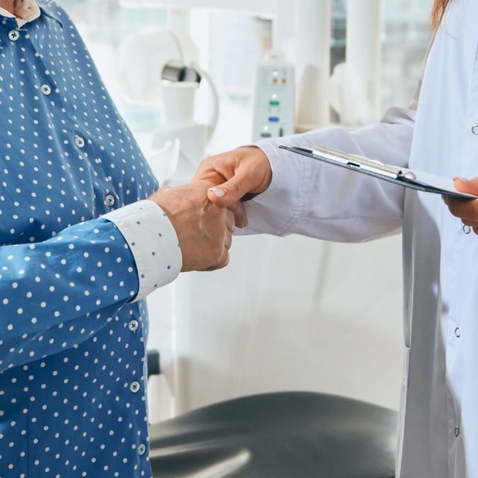 A person in a blue polka-dot shirt shakes hands with a person in a white coat, holding a clipboard, in a medical setting.