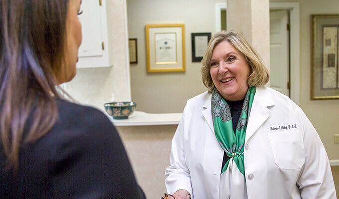 Doctor in white coat smiles, talking with a patient. Artwork and a doorway are visible in the background.