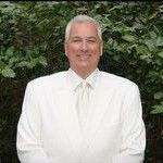 A man in a white shirt and tie is standing in front of a bush.