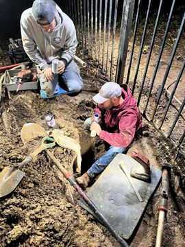 Two people digging in a dirt pit beside a fence. One kneels holding a light, the other kneels looking on. Tools are visible.