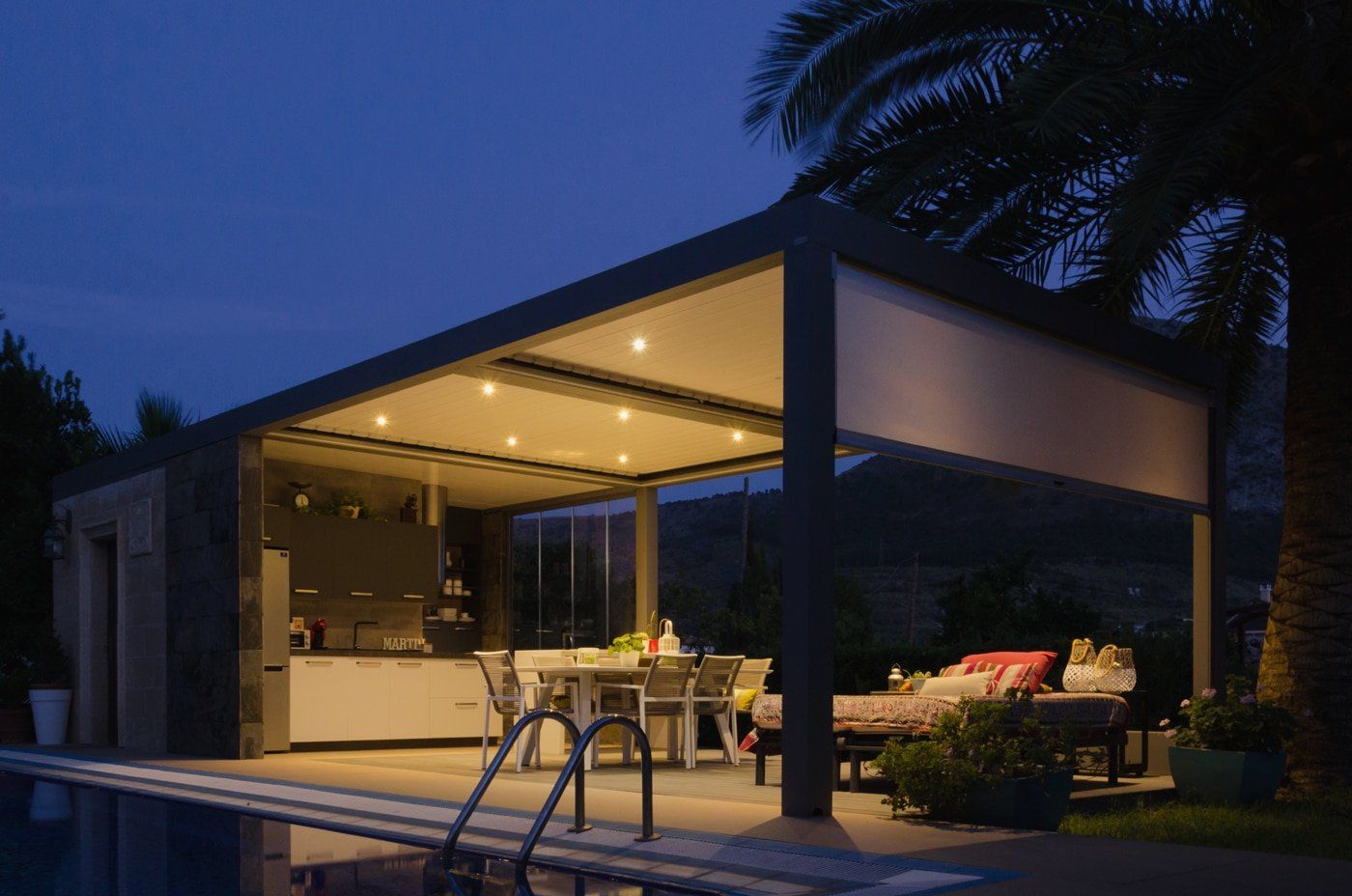 Un patio con pérgola con vistas a una piscina por la noche.