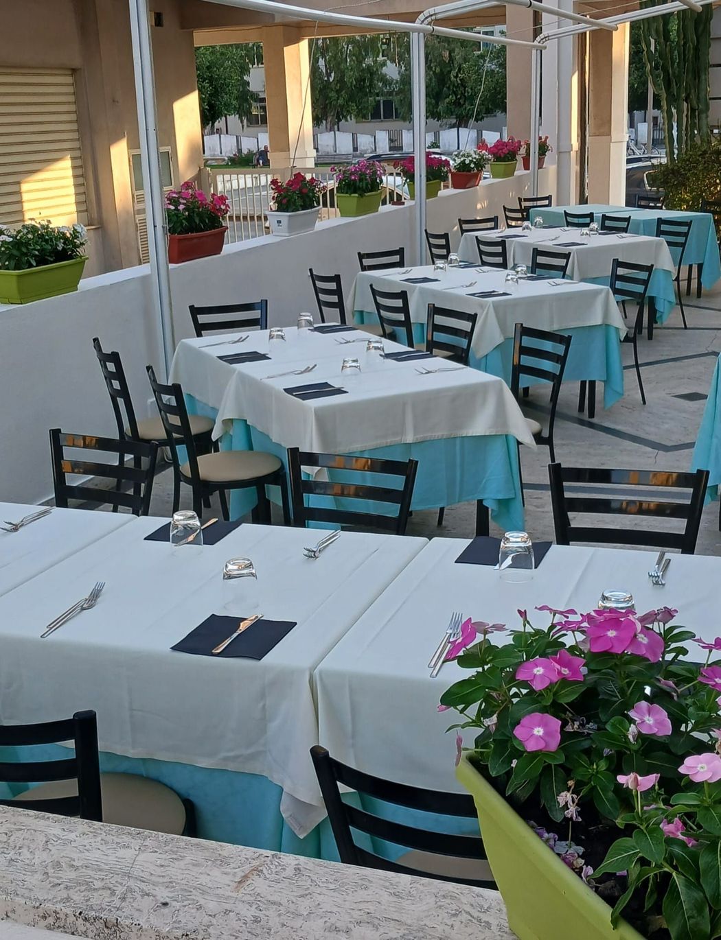 Patio esterno con tavoli ricoperti di tovaglie bianche, sedie turchesi e fiori rosa vicino all'ingresso di un ristorante.