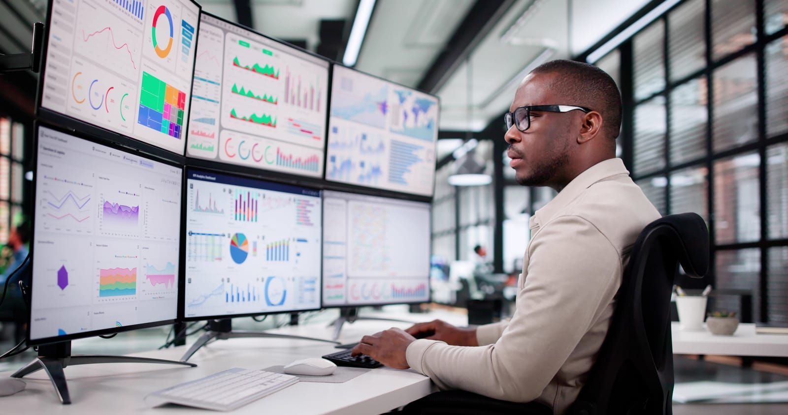 A man is sitting at a desk in front of a bunch of computer monitors looking at the marketing trends.