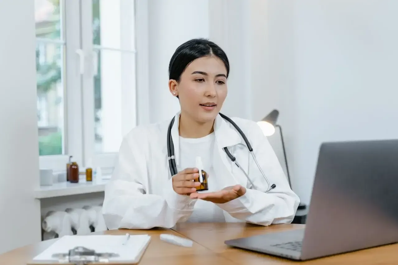 Doctor in a white coat speaking on a video call at a desk with a laptop, holding a cup in a bright office.
