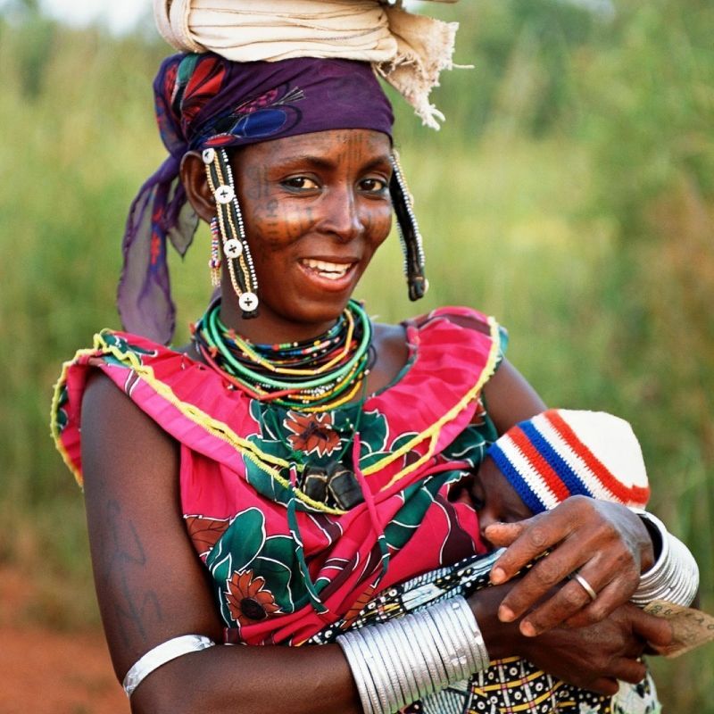A Fulani woman in Benin, dressed in vibrant traditional attire, carries a baby in a sling and balances a bucket on her head—a striking image of daily life and cultural pride. The Fulani are known for their nomadic heritage, colorful clothing, and strong community traditions. This scene reflects the rhythm and resilience of rural Benin, where heritage and modern life coexist. Inspiration Africa organizes authentic journeys to Benin, offering opportunities to engage respectfully with local cultures, explore Fulani traditions, and experience the richness of West African life through immersive, tailor-made travel experiences designed around your interests.