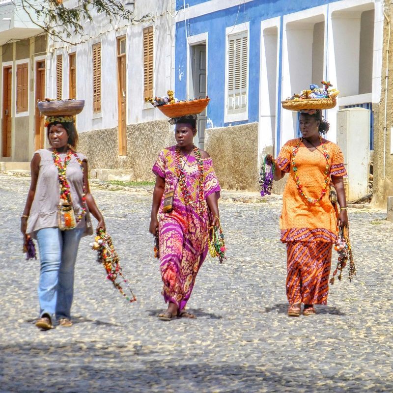 Three women walk down a sunlit street in Cape Verde, balancing baskets on their heads and wearing vibrant traditional dresses—a daily scene full of rhythm, resilience, and island charm. Cape Verde’s culture blends African, Portuguese, and Creole influences, best experienced through its people, markets, and music-filled neighborhoods. Inspiration Africa designs custom journeys to Cape Verde, connecting travelers with authentic local life across the islands. Whether exploring cobbled streets, coastal villages, or cultural traditions, we’ll take you beyond the expected. Travel with us to discover the true spirit of Cape Verde—alive in every step, smile, and color.