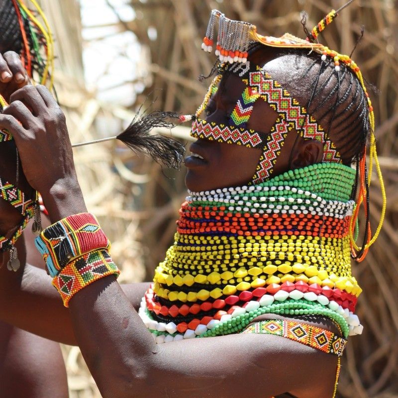 Capture the intimate dynamics of the Turkana Tribe in Kenya with this unique photo. A woman, adorned in beautiful traditional attire and jewelry, carefully adjusts or helps put on a man's clothing. The man respectfully has his head bowed, highlighting the communal roles and traditions of the Turkana people. Inspiration Africa specializes in designing your unique, tailor-made trip to Kenya for profound cultural encounters and exploring other spectacular destinations across Africa. Discover authentic Northern Kenya with us!