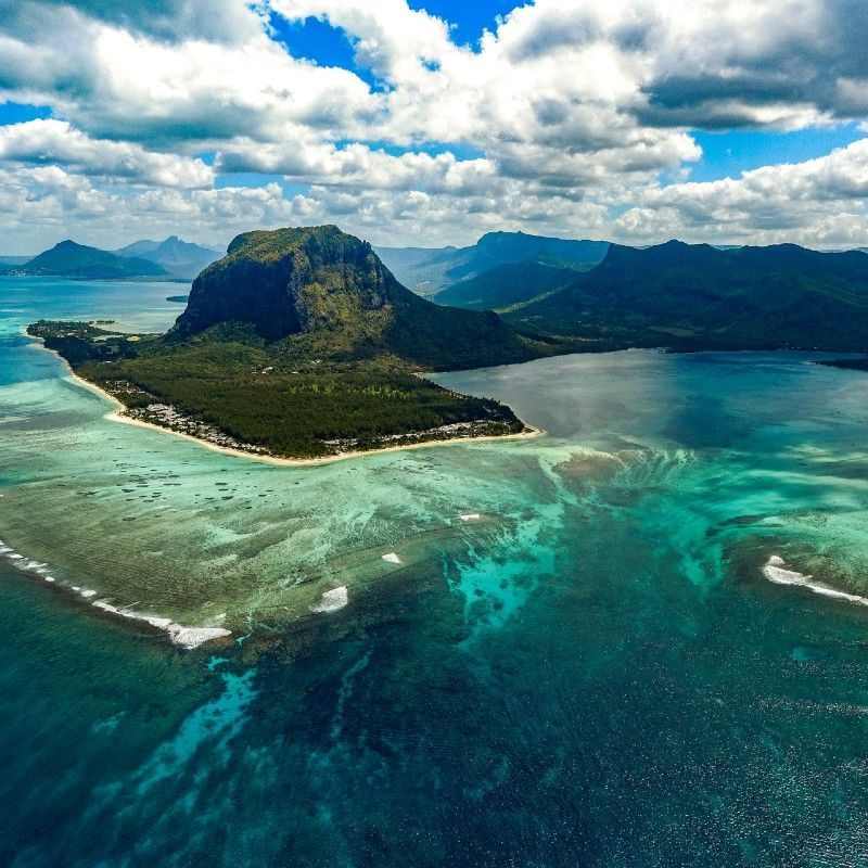 An aerial view of Le Morne reveals its dramatic peak surrounded by turquoise waters, coral reefs, and the breathtaking illusion of an underwater waterfall—a mesmerizing natural wonder off the coast of Mauritius.