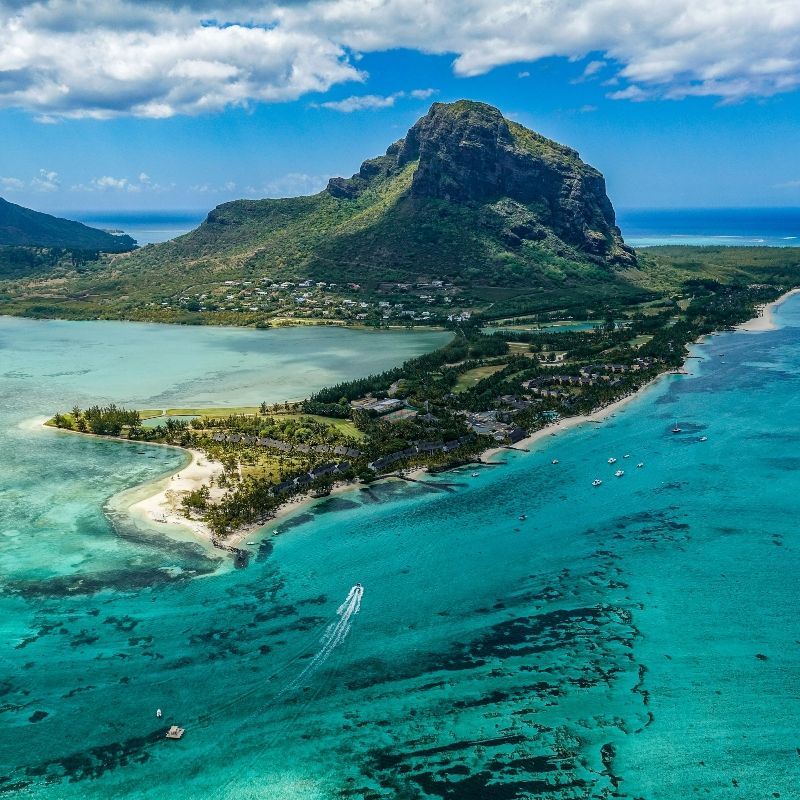 Witness the awe-inspiring Le Morne Underwater Waterfall in Mauritius from the air. This aerial shot captures the mesmerizing optical effect, where varying shades of ocean water—from deep sapphire blue to bright turquoise—create the illusion of a flowing cascade. The majestic Le Morne mountain, draped in lush green vegetation, dramatically anchors the scene. Inspiration Africa designs your ultimate, tailor-made trip to Mauritius and other exceptional African destinations. Explore this geological wonder and book your dream island getaway with us!