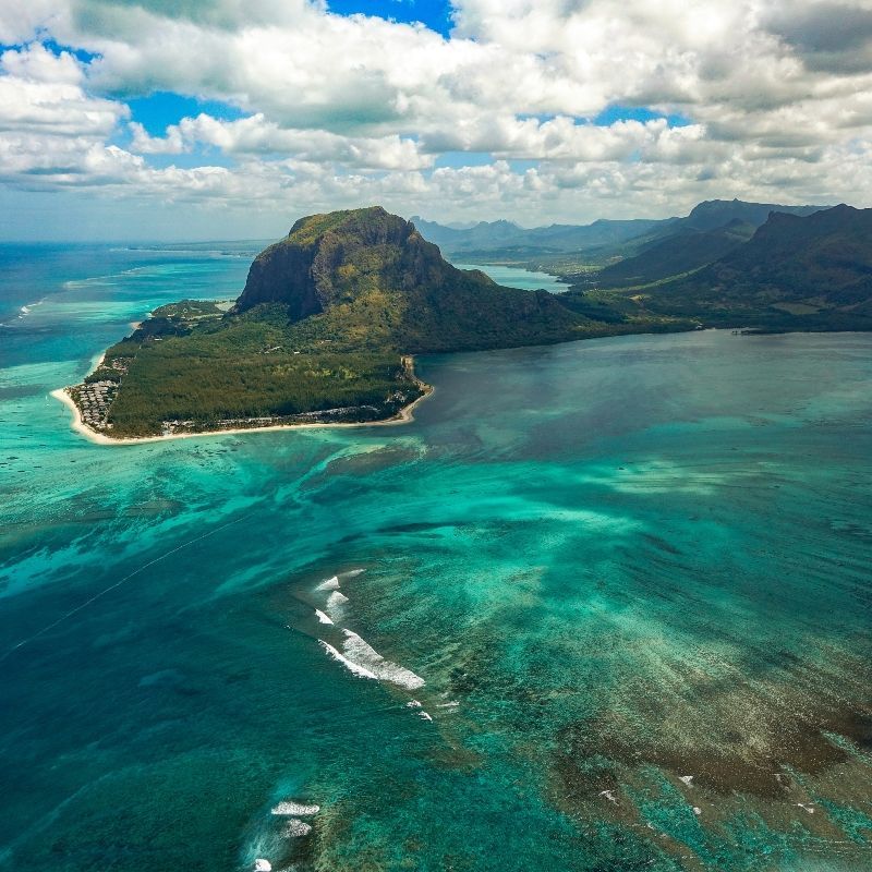 Witness the awe-inspiring Le Morne Underwater Waterfall in Mauritius from the air. This aerial shot captures the mesmerizing optical effect, where varying shades of ocean water—from deep sapphire blue to bright turquoise—create the illusion of a flowing cascade. The majestic Le Morne mountain, draped in lush green vegetation, dramatically anchors the scene. Inspiration Africa designs your ultimate, tailor-made trip to Mauritius and other exceptional African destinations. Explore this geological wonder and book your dream island getaway with us!