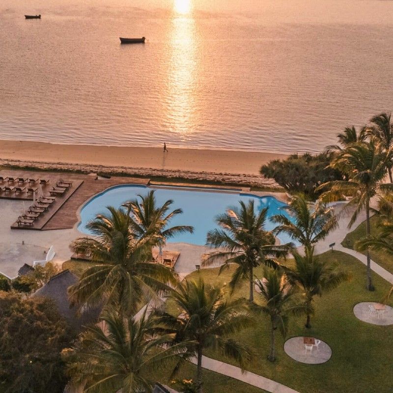 The pool area at Vilanculos Beach Lodge in Mozambique is captured in this breathtaking aerial travel photograph at sunset, showcasing an infinity-style pool that overlooks the sparkling Indian Ocean. The pool reflects the warm hues of the setting sun—gold, pink, and purple—while sun loungers and shaded areas surround the water, inviting guests to relax in a tranquil, luxurious setting. Lush tropical gardens and the coastline frame the scene, creating a seamless connection between the lodge, the beach, and the sea. The photograph conveys serenity, elegance, and the magic of Mozambique’s coastal beauty at twilight, highlighting Vilanculos Beach Lodge as an idyllic retreat where ocean vistas and sunset panoramas define the experience. Inspiration Africa specializes in bespoke, tailor-made journeys to Mozambique and across Africa, crafting unforgettable travel experiences in extraordinary destinations.
