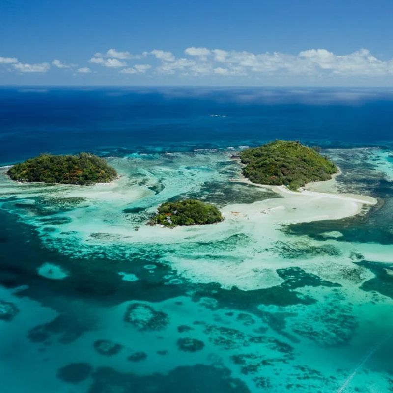 An aerial view captures three small islands in Sainte Anne Marine National Park in the Seychelles, showcasing powder-white sand beaches, lush green vegetation, and stunning turquoise ocean waters in multiple shades of blue. Surrounded by protected coral reefs and crystal-clear seas, this pristine marine sanctuary highlights the natural beauty and biodiversity of the Indian Ocean. The tranquil islands offer exceptional snorkeling, kayaking, and island-hopping experiences in an unspoiled tropical setting. Ideal for nature lovers, beach enthusiasts, and photographers, this scene embodies barefoot luxury and island serenity. Inspiration Africa specializes in bespoke, tailor-made journeys to the Seychelles and across Africa, crafting exclusive travel experiences that reveal extraordinary island landscapes, marine wonders, and unforgettable coastal escapes.
