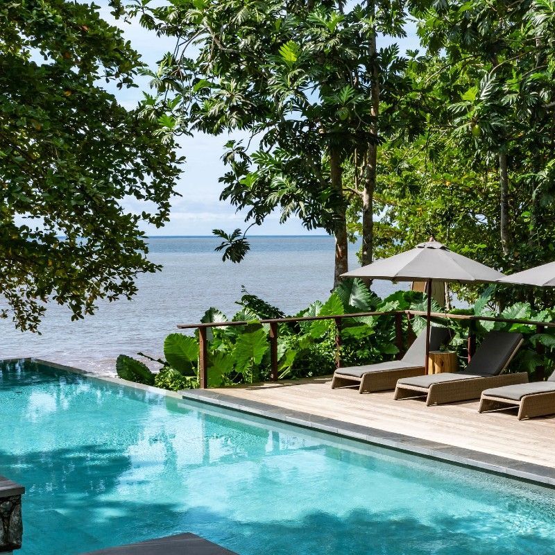 The oceanfront swimming pool at Sundy Praia in São Tomé and Príncipe is captured in this striking travel photograph, showcasing a secluded luxury retreat set between lush tropical forest and the Atlantic Ocean. The pool blends seamlessly into the natural landscape, framed by palm trees, soft sandy tones, and verdant greenery, with uninterrupted views toward the sea beyond. Sun loungers and shaded areas invite quiet relaxation, while the design reflects a balance of understated elegance and barefoot luxury. The scene conveys a sense of tranquility, exclusivity, and connection to nature, highlighting Sundy Praia as an idyllic island escape. Inspiration Africa specializes in bespoke, tailor-made journeys to São Tomé and Príncipe and across Africa, crafting unforgettable travel experiences in extraordinary destinations.
