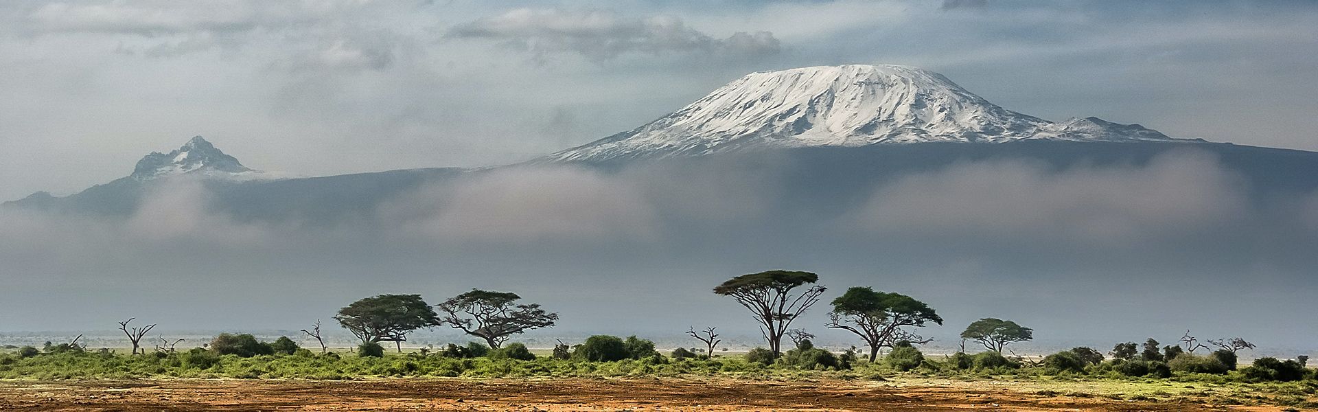 Mount Kilimanjaro rises majestically above the clouds, its snowy peak contrasting sharply with the vast savannah stretching below. The iconic mountain stands as a serene, towering presence over the African landscape.