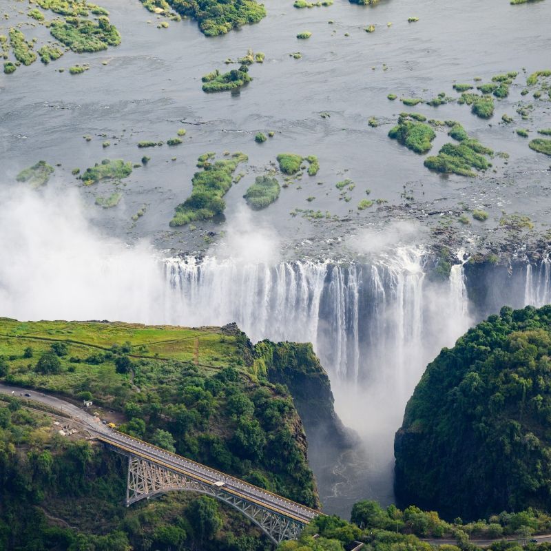 Aerial views of Victoria Falls in Zimbabwe reveal cascading water plummeting into mist-filled gorges, surrounded by lush landscapes and the dramatic Zambezi River.