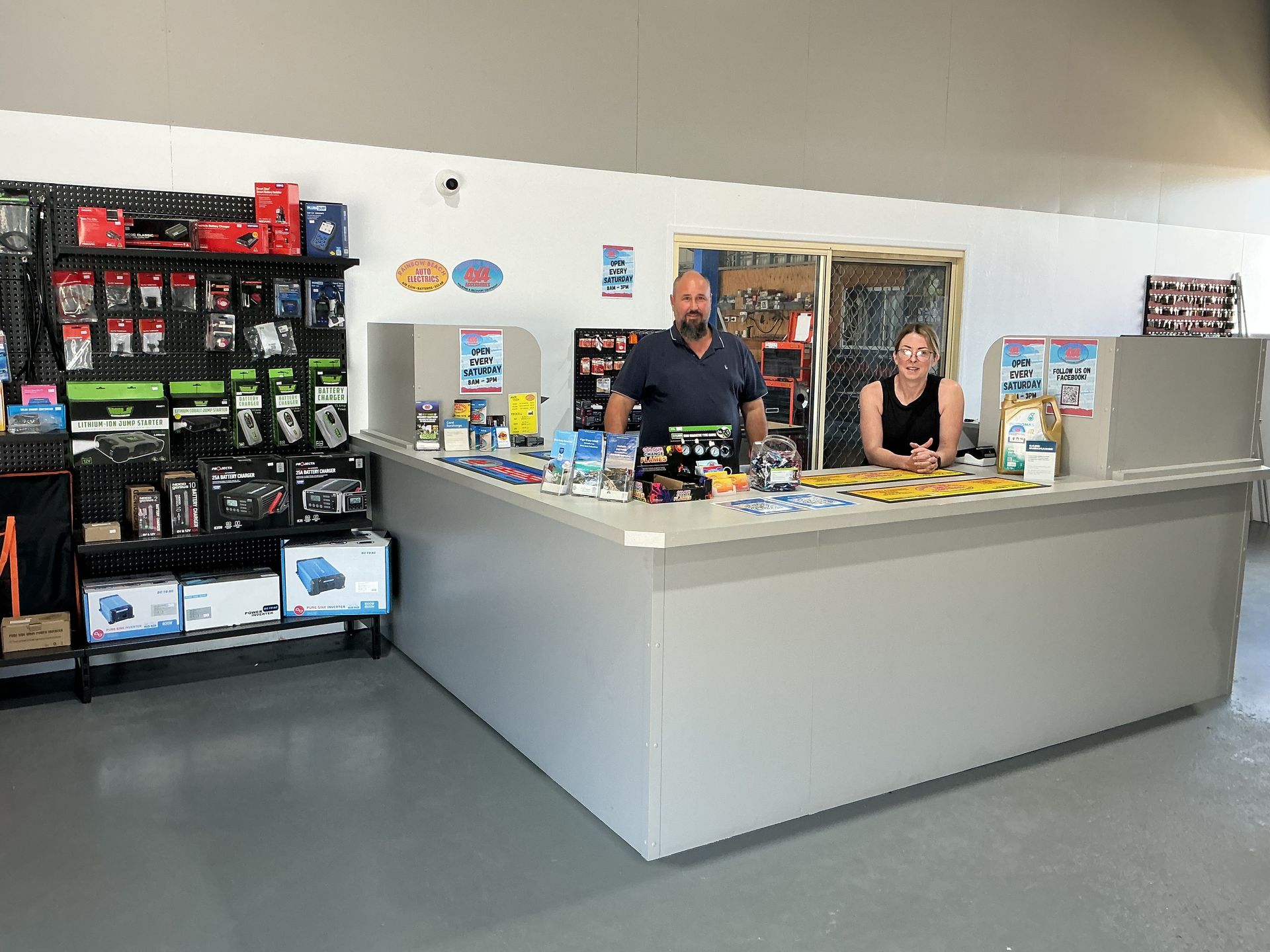 Two people behind a gray counter in a store. Shelves with products on left. Gray floor, white walls. — Rainbow Beach 4x4 Accessories in Rainbow Beach, QLD