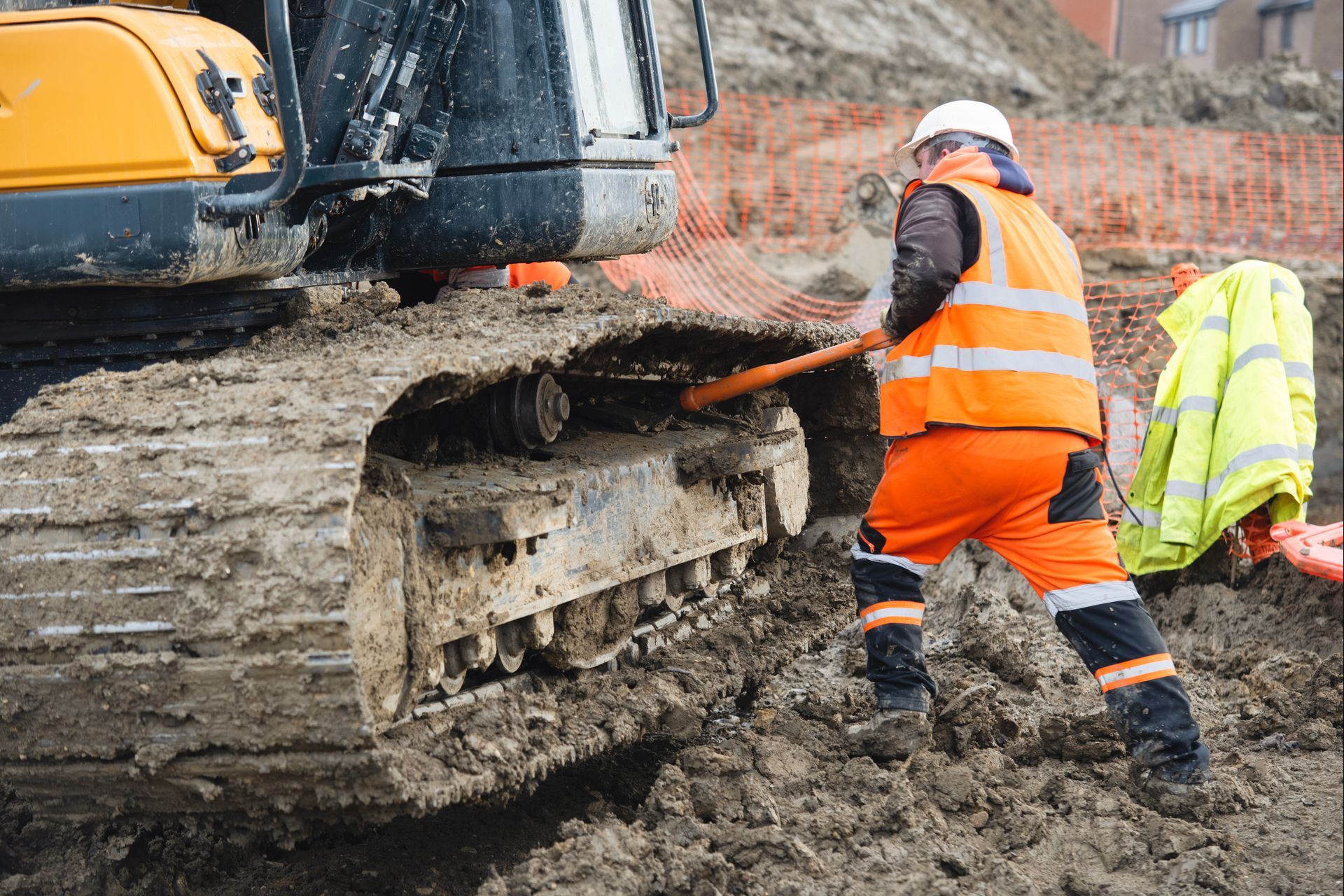 Construction worker clearing mud from the track of an excavator with a long-handled tool.