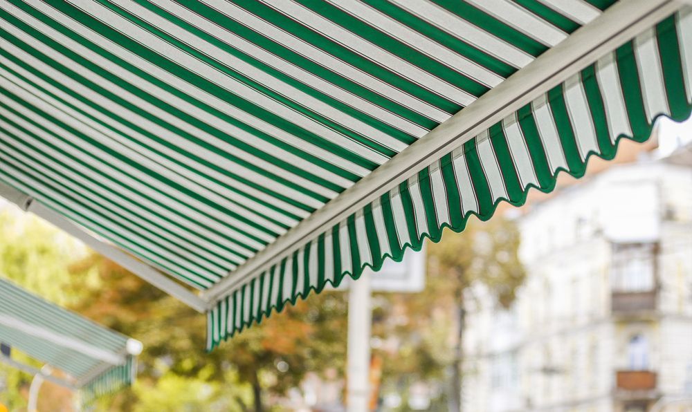 A Green and White Striped Awning is Hanging From the Roof of a Building — Elite Window Furnishings In Woden, ACT
