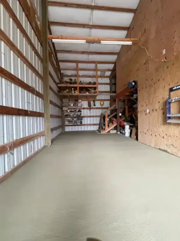 A freshly poured concrete floor inside a large workshop with metal siding and wood shelving.