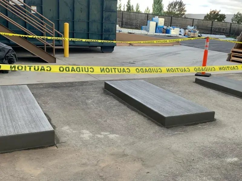 Three gray concrete pads, likely for waste containers, surrounded by caution tape in an outdoor commercial setting.