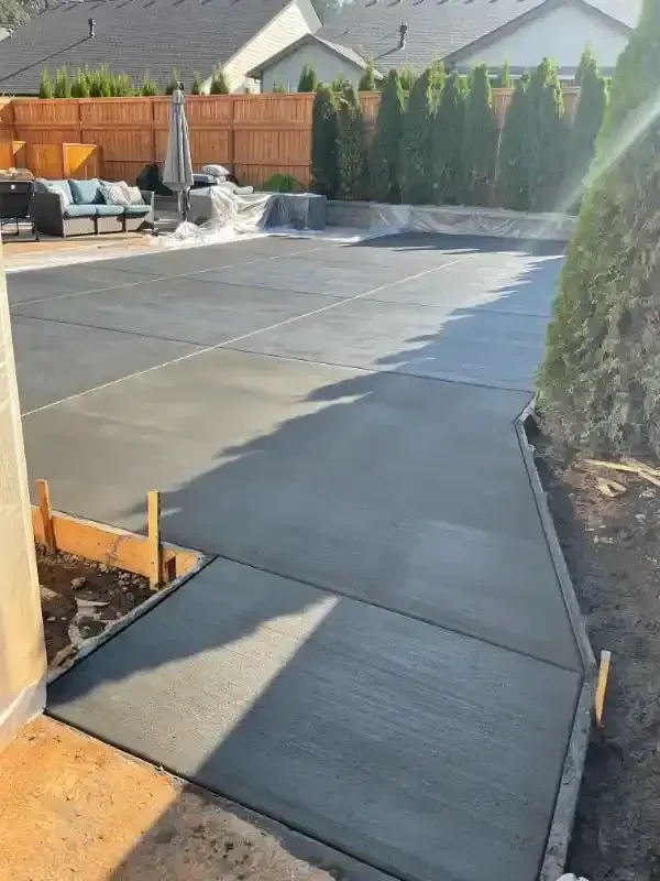 Freshly poured concrete pathway leading to a backyard patio with seating and tall green hedges.