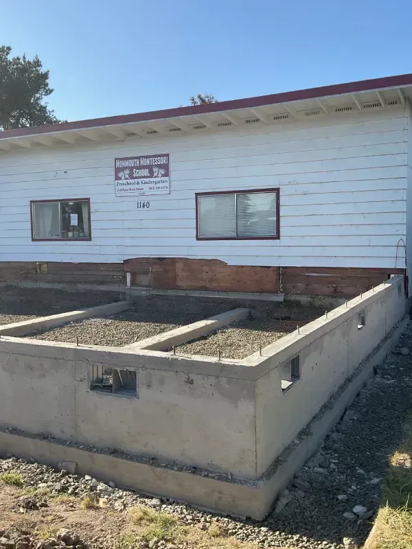 Building on a concrete foundation, white exterior, sign, two windows, clear sky.