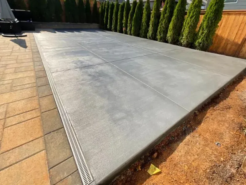 Concrete patio with drainage, adjacent to a paver patio, lined by evergreen trees and a wood fence.