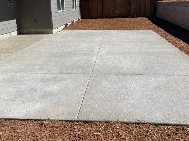 Concrete patio with joint lines surrounded by red mulch; backdrop of a building and a wooden fence.