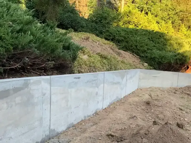 Gray concrete retaining wall with dirt in front, lush green hillside behind.