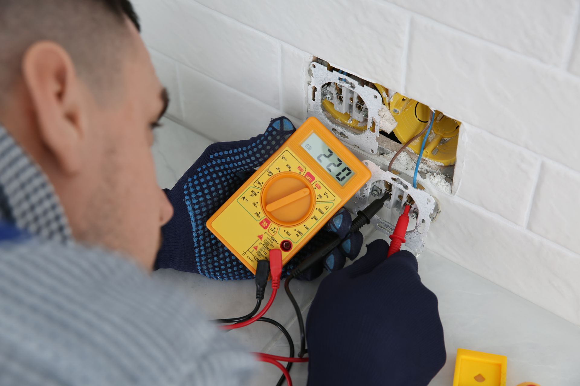 Technician using a yellow multimeter to test exposed wiring in a white tiled wall outlet box