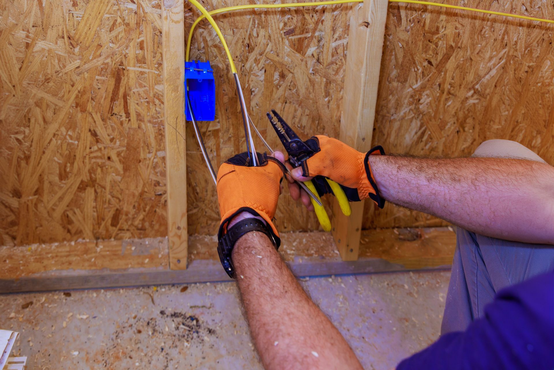 Gloved hands cutting electrical wires inside a wall cavity with pliers and a blue outlet box
