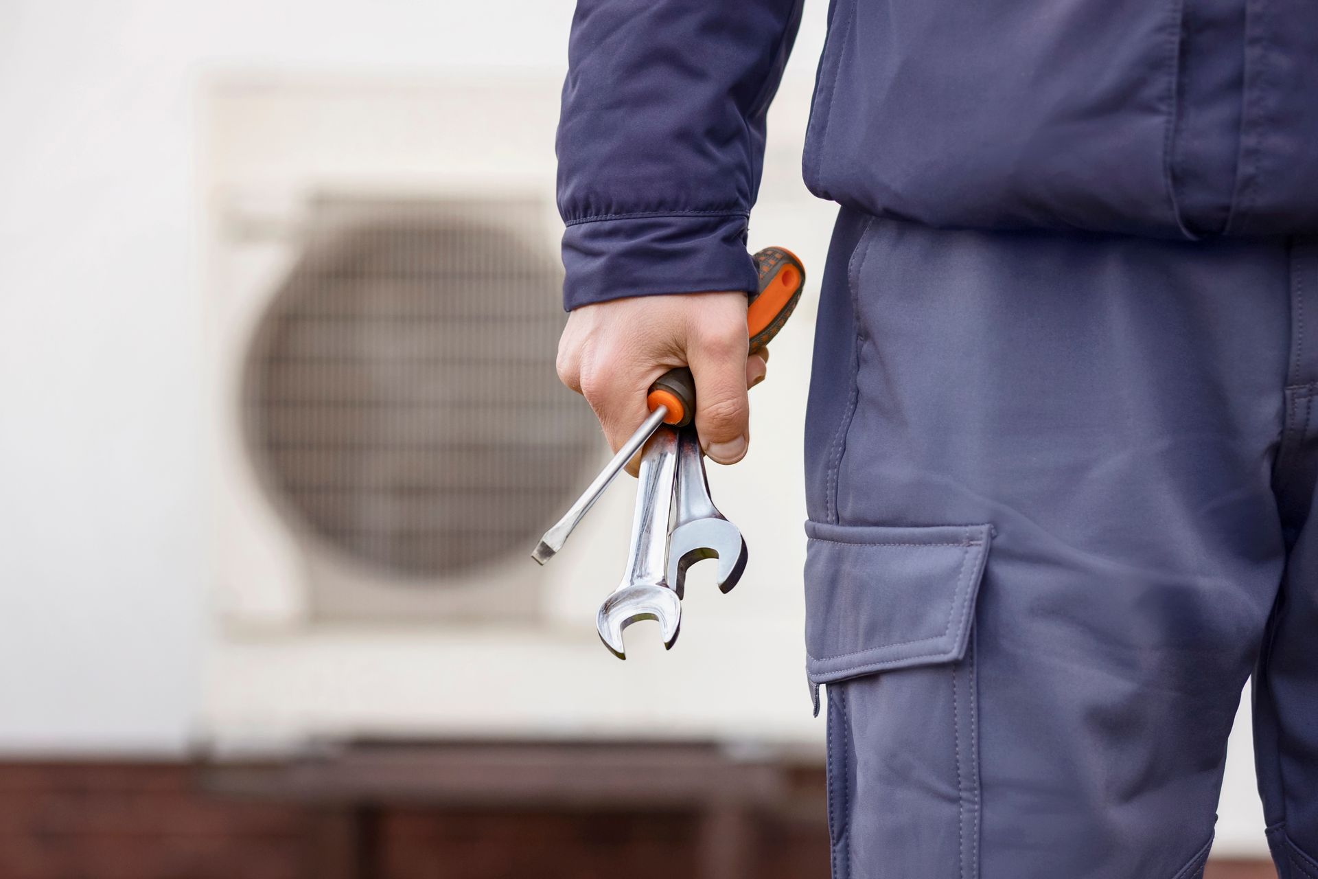 Person in blue work clothes holding wrench and pliers beside an outdoor air conditioner unit