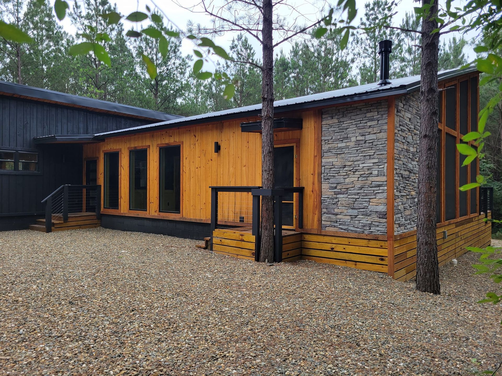 A wooden house with a stone wall is surrounded by gravel and trees.