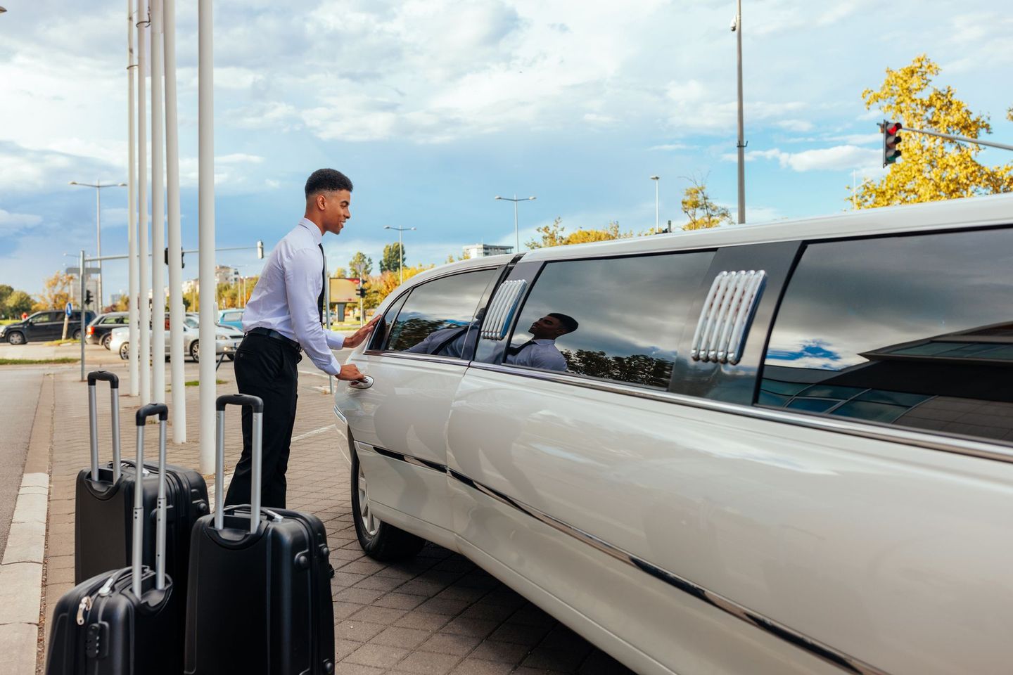 Man opening door of white limousine, two suitcases next to the car, outdoor setting with poles, and a cloudy sky.