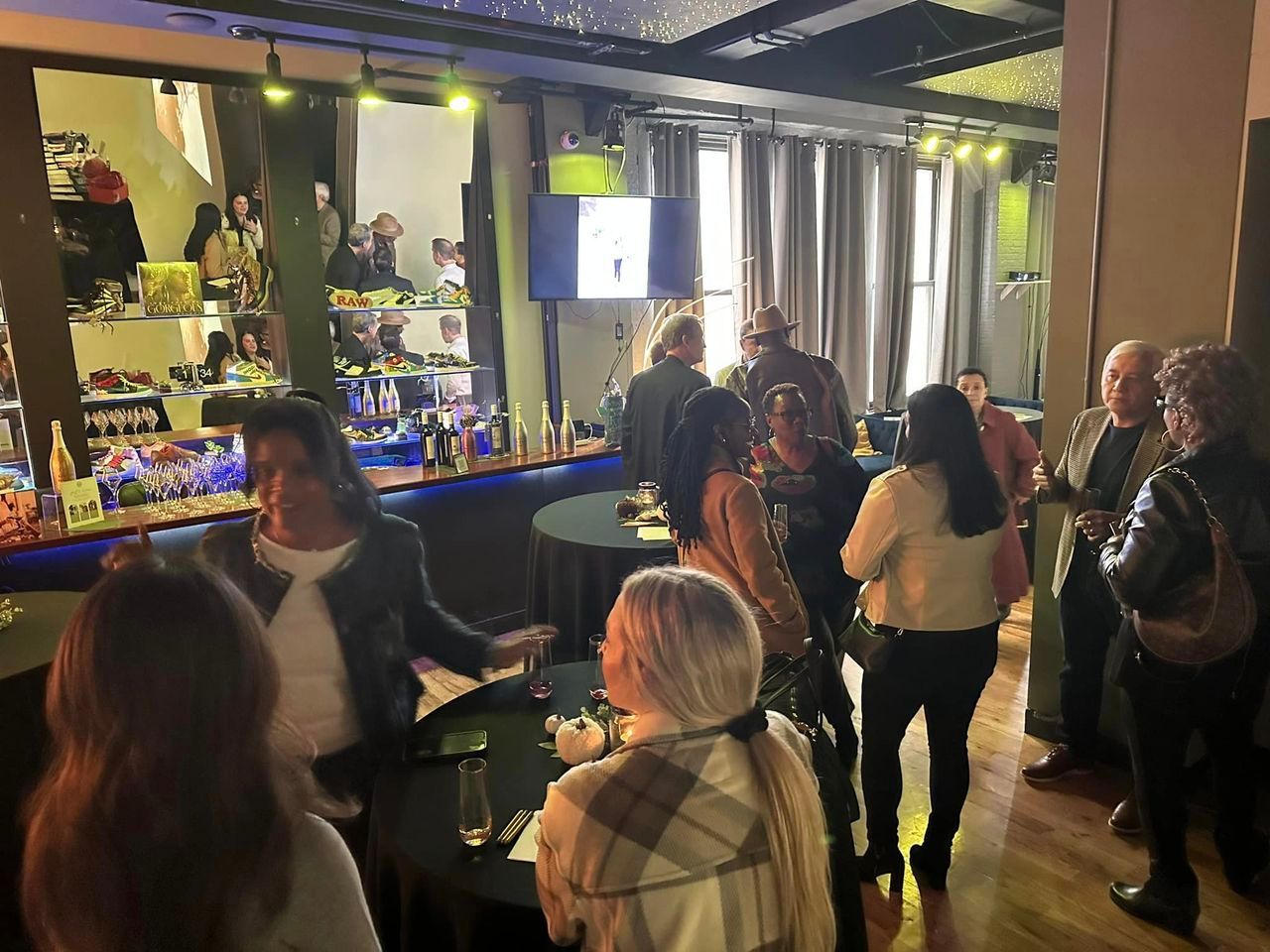 People mingling at a bar, some near the counter. Dim lighting, drinks visible, warm tones.
