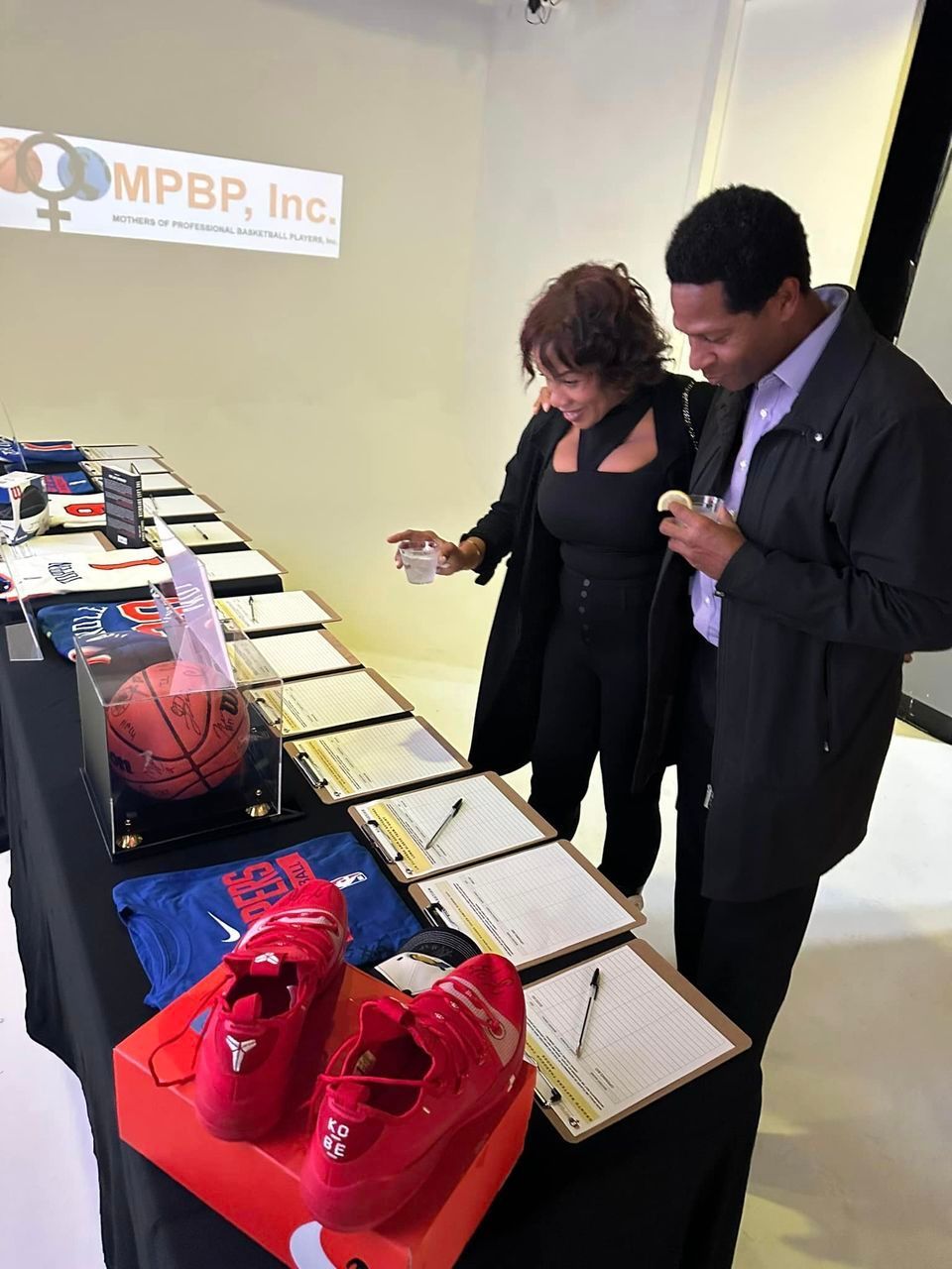 Two people looking at items on a table, likely at an auction. Basketball and shoes are visible.