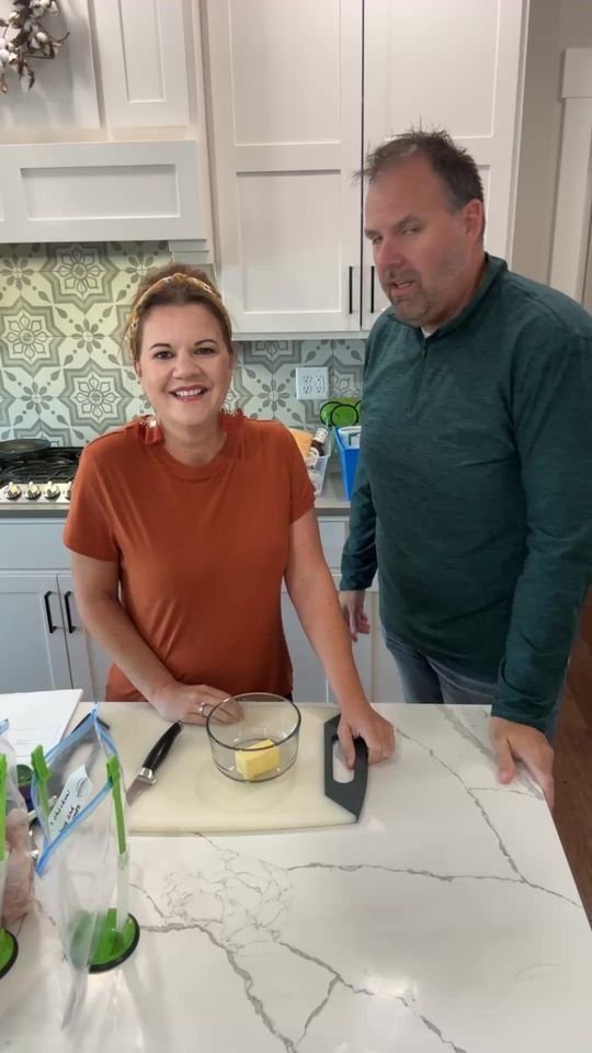 Woman and man in kitchen, woman smiles, cutting butter, man watches.