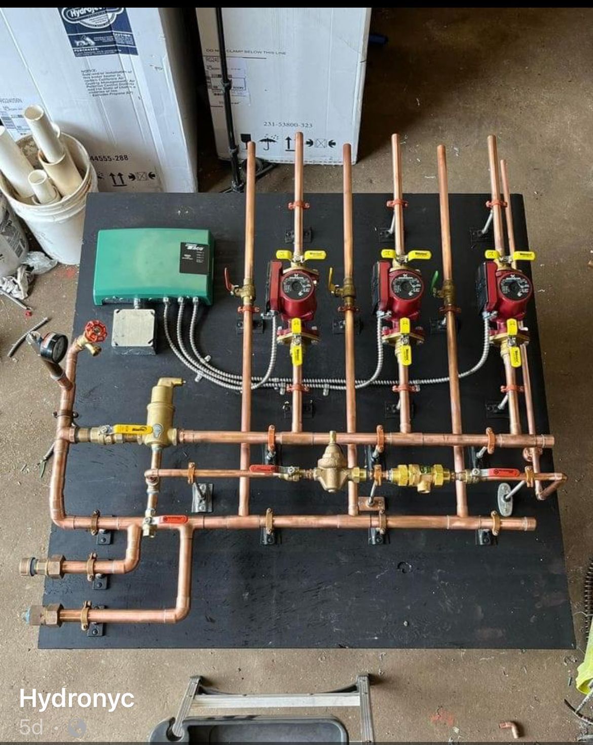 Copper hydronic plumbing manifold with three red circulator pumps on a black board, labeled “Hydronic”