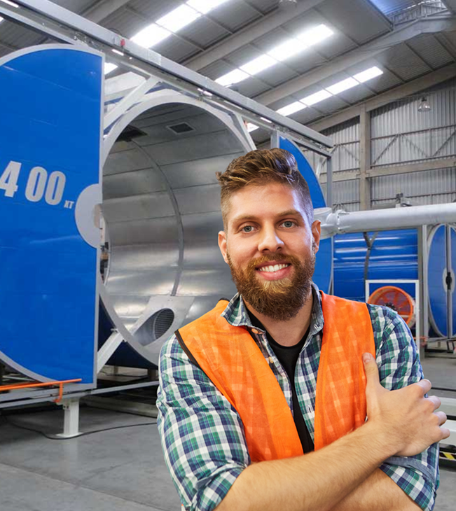 Man in safety vest smiles in a factory setting, with blue and silver industrial equipment in background.
