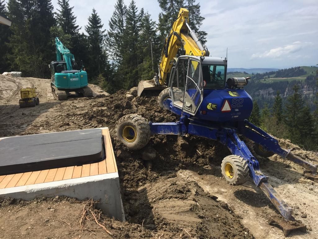 Baustelle mit zwei Baggern, die Erde in der Nähe eines Betonbauwerks bewegen; im Hintergrund ein bewaldeter Hang.