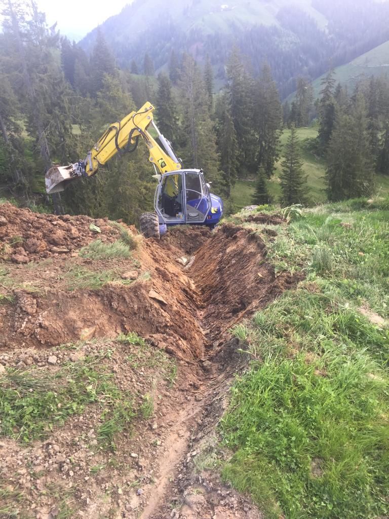 Ein Bagger gräbt einen Graben in einem grasbewachsenen, abschüssigen Feld in der Nähe von Bäumen und Bergen.