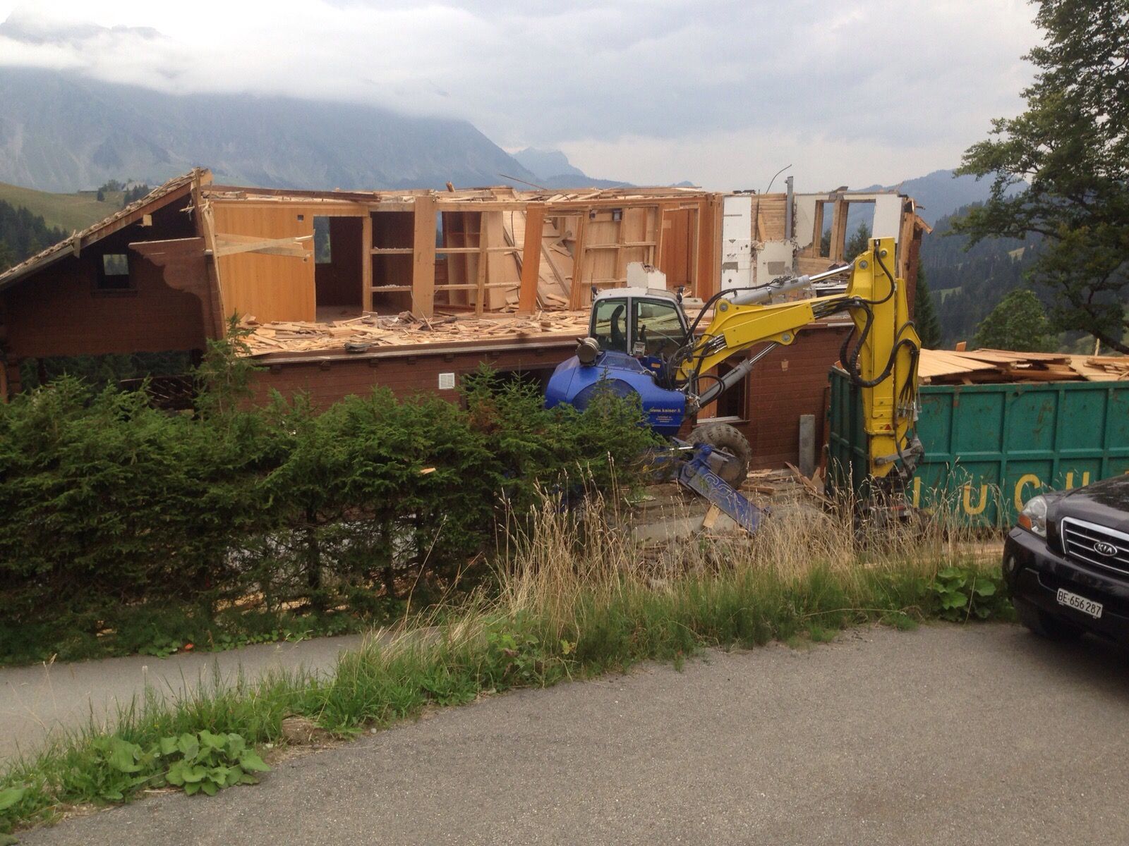 Ein Bagger reißt ein Holzgebäude in einer Berglandschaft ab. Trümmer und ein grüner Müllcontainer sind zu sehen.
