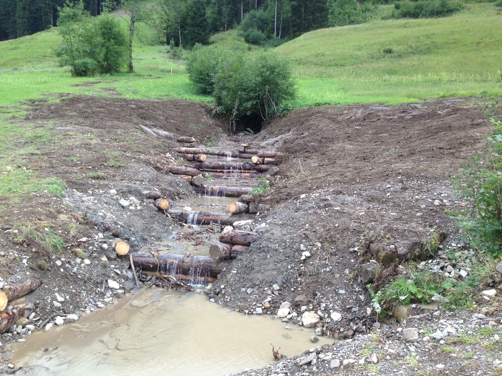 Ein künstlich angelegter Bachlauf aus Baumstämmen bildet einen kleinen Wasserfall in einer grasbewachsenen Hanglandschaft.
